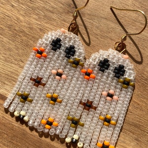 K&ouml;nnte beinhalten: Ein Paar wei&szlig;e Perlenohrringe mit einem geometrischen Muster in Orange, Schwarz, Braun und Creme. Die Ohrringe sind aus kleinen Perlen gefertigt und haben goldene Haken.