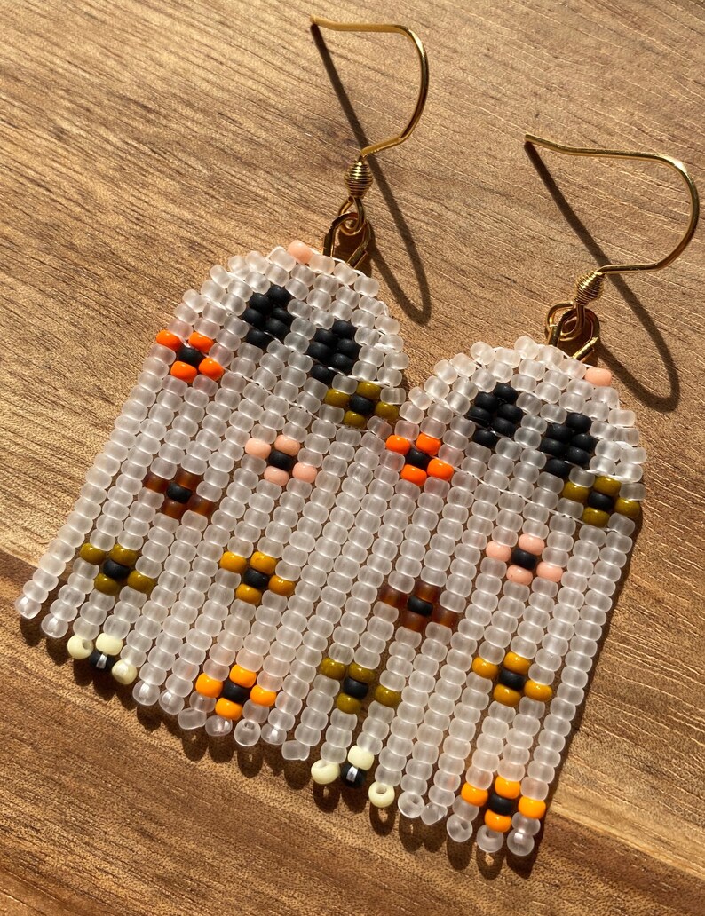 K&ouml;nnte beinhalten: Ein Paar wei&szlig;e Perlenohrringe mit einem geometrischen Muster in Orange, Schwarz, Braun und Creme. Die Ohrringe sind aus kleinen Perlen gefertigt und haben goldene Haken.