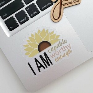 May include: A clear sticker with a yellow sunflower and the text "I AM capable worthy enough" in black, pink, and brown lettering.