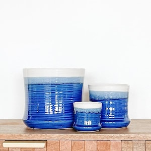 May include: Three blue ceramic planters with white rims. The planters are of different sizes and are arranged on a wooden surface.