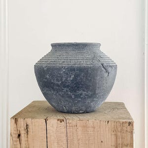 May include: A dark grey, textured vase with a ridged neck sits on a weathered, light-coloured wooden pedestal. The vase has a rounded body and a slightly rough surface. The pedestal is a square block of wood with visible grain and knots.