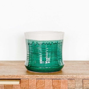 May include: A green glass candle jar with a white top sits on a wooden table with woven wall decor behind it.