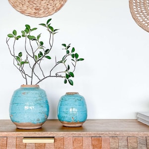 May include: A wooden dresser with two drawers and a set of six woven wall decorations in various shapes and sizes. Two turquoise ceramic vases with a distressed finish are on top of the dresser, one with a small leafy branch inside.