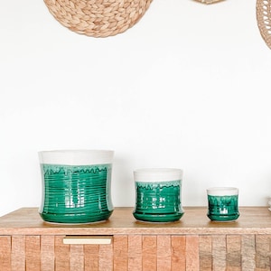 May include: Three green ceramic planters with white rims, sitting on a wooden shelf. The planters are different sizes and have a textured, striped design.