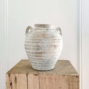 May include: A distressed white and beige ceramic vase with two handles is displayed on a weathered wooden block. The vase has a textured, aged look with horizontal lines. The wooden block shows visible grain and knots, giving a rustic feel.