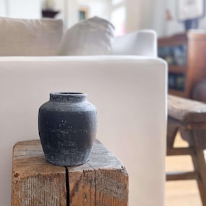 Peut inclure: Un petit vase gris foncé à surface texturée est posé sur un bloc de bois rectangulaire vieilli. Le vase a un corps arrondi et un col étroit. Le bloc de bois présente un grain visible et un aspect naturel et rustique. L'arrière-plan est un salon flou.