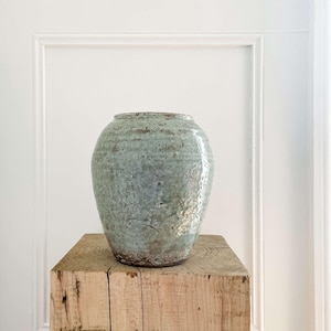 May include: A light blue ceramic vase with a textured surface and rounded form rests on a weathered, square wooden pedestal. The vase has a glossy finish and a mottled appearance. The pedestal is made of rough-hewn wood, showing visible grain and knots.
