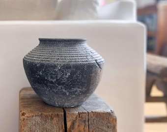 Könnte beinhalten: Eine dunkelgraue, strukturierte Keramikvase mit rundem Körper und strukturiertem Rand. Die Vase steht auf einem verwitterten Holzblock. Der Hintergrund zeigt ein weißes Sofa und einen Holzstuhl, was auf Wohnkultur hindeutet.