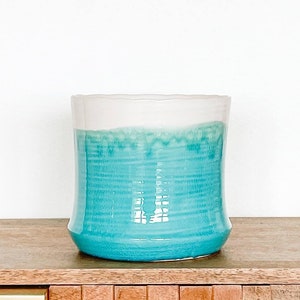 May include: A teal blue ceramic vase with a white rim. The vase is sitting on a wooden surface.