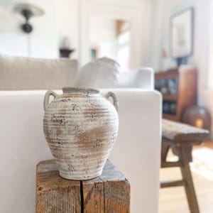 K&ouml;nnte beinhalten: Eine dekorative Vase in Off-White und Braun mit zwei Griffen. Die Vase hat eine strukturierte, verwitterte Oberfl&auml;che und steht auf einem verwitterten Holzblock. Der Hintergrund zeigt ein wei&szlig;es Sofa und eine Holzbank, was auf ein Wohn-Dekor-Thema hindeutet.