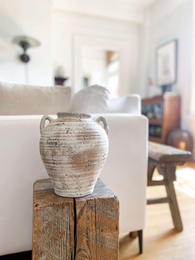 K&ouml;nnte beinhalten: Eine dekorative Vase in Off-White und Braun mit zwei Griffen. Die Vase hat eine strukturierte, verwitterte Oberfl&auml;che und steht auf einem verwitterten Holzblock. Der Hintergrund zeigt ein wei&szlig;es Sofa und eine Holzbank, was auf ein Wohn-Dekor-Thema hindeutet.