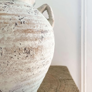 K&ouml;nnte beinhalten: Eine gro&szlig;e, cremefarbene Keramikvase mit zwei Henkeln. Die Vase hat eine strukturierte, abgenutzte Oberfl&auml;che mit braunen und schwarzen Akzenten. Die Vase steht auf einer verwitterten Holzoberfl&auml;che.