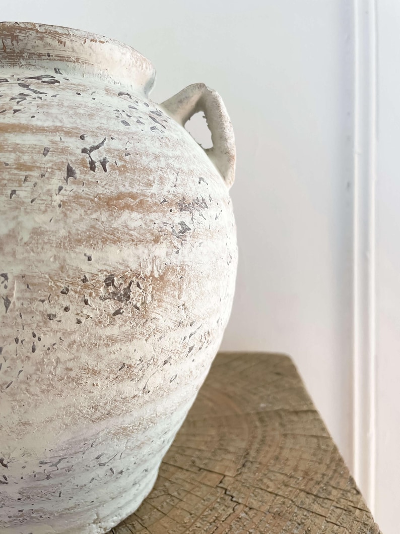 K&ouml;nnte beinhalten: Eine gro&szlig;e, cremefarbene Keramikvase mit zwei Henkeln. Die Vase hat eine strukturierte, abgenutzte Oberfl&auml;che mit braunen und schwarzen Akzenten. Die Vase steht auf einer verwitterten Holzoberfl&auml;che.