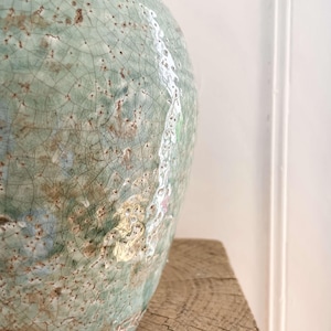 May include: A large, round ceramic vase with a crackled, celadon glaze. The vase has a textured surface with brown and white accents. The vase is sitting on a rough-hewn wooden surface. The overall aesthetic is rustic and artisanal.