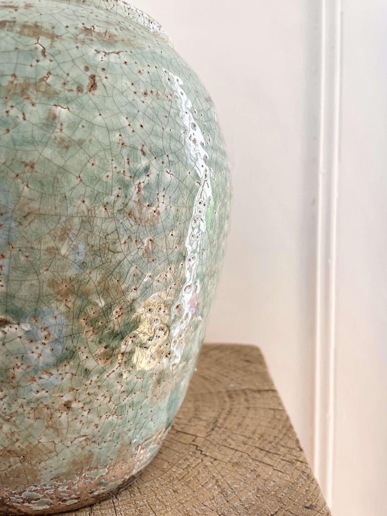 May include: A large, round ceramic vase with a crackled, celadon glaze. The vase has a textured surface with brown and white accents. The vase is sitting on a rough-hewn wooden surface. The overall aesthetic is rustic and artisanal.