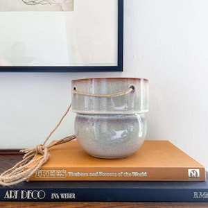 May include: A two-part ceramic hanging planter with a light green and brown glaze. The top section has a brown rim and holes for a rope. The planter is resting on a stack of books, including one titled "TREES Timbers and Forests of the World."