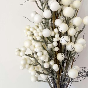 以下が含まれることがあります： 白い球状のベリーが付いた装飾用の人工植物のクローズアップ。ベリーはサイズが異なり、細い茶色と灰色の枝に集まっています。背景は無地の白です。