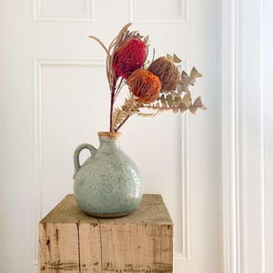 May include: A pale blue ceramic vase with a handle, containing dried flowers in red, orange, and brown hues. The vase rests on a rustic wooden block, creating a natural aesthetic. The background features a white door.