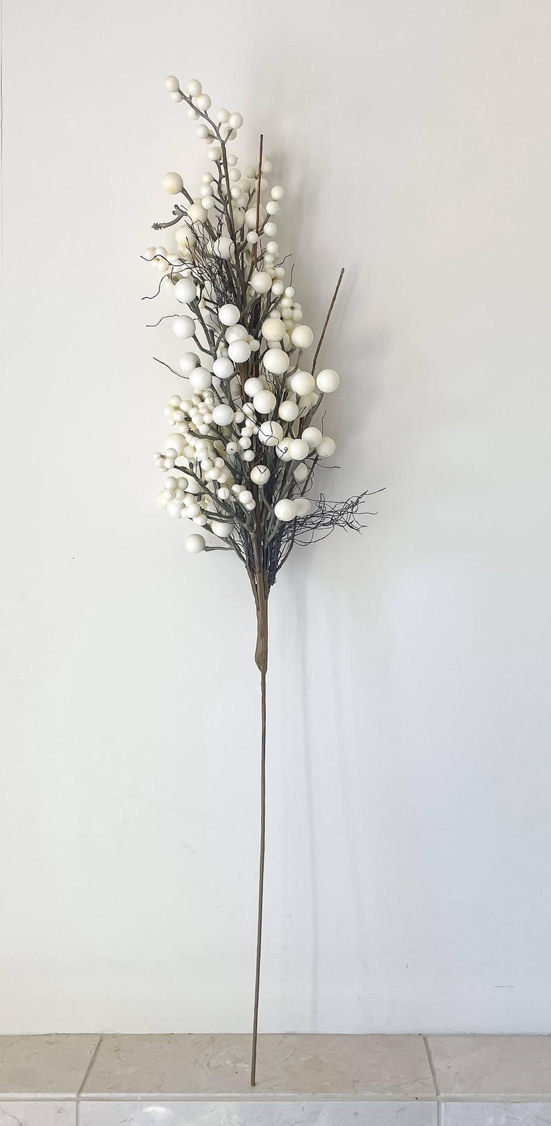 以下が含まれることがあります： 白い球形のベリーの房が付いた、人工的な花の茎。 ベリーは、暗い小枝のような枝に密集しており、長く細い茶色の茎が付いています。 背景はプレーンな白い壁です。