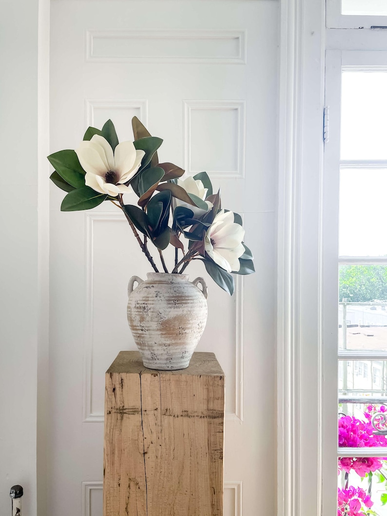 K&ouml;nnte beinhalten: Eine wei&szlig; get&uuml;nchte Vase mit zwei Griffen enth&auml;lt mehrere gro&szlig;e wei&szlig;e Magnolienbl&uuml;ten mit dunkelgr&uuml;nen Bl&auml;ttern. Die Vase steht auf einem verwitterten, hellen Holzsockel vor einer wei&szlig;en T&uuml;r.