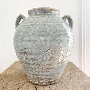 Peut inclure: Un grand vase en céramique vieillie avec deux anses. Le vase présente un glaçage craquelé bleu-gris clair et repose sur un bloc de bois rustique. L'aspect vieilli suggère une esthétique vintage ou antique, adaptée à la décoration intérieure.