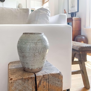 May include: A weathered, light green ceramic vase with a textured surface sits atop a rustic, square wooden block. The vase has a rounded shape and a slightly flared rim. The wood block displays visible grain and a natural, aged appearance. The background includes a white sofa and a wooden stool.