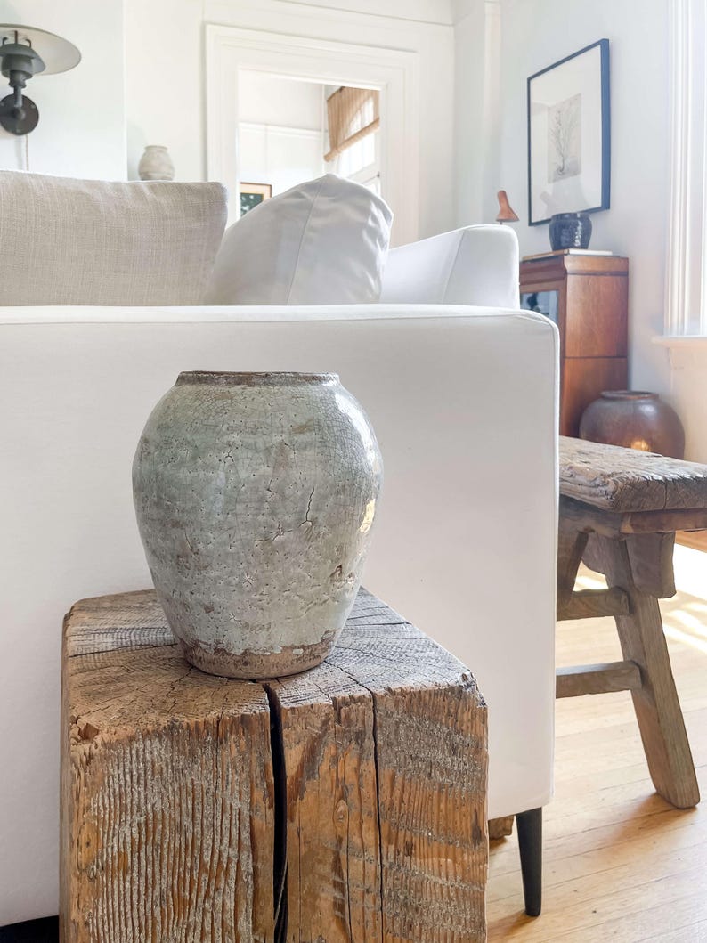 May include: A weathered, light green ceramic vase with a textured surface sits atop a rustic, square wooden block. The vase has a rounded shape and a slightly flared rim. The wood block displays visible grain and a natural, aged appearance. The background includes a white sofa and a wooden stool.