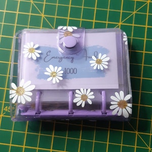 Puede incluir: Un archivador de plástico transparente con un mecanismo de anillas morado y una tarjeta blanca en el interior. La tarjeta tiene el texto "Emergency Fund 1000" y está decorada con margaritas blancas con centros de purpurina dorada.