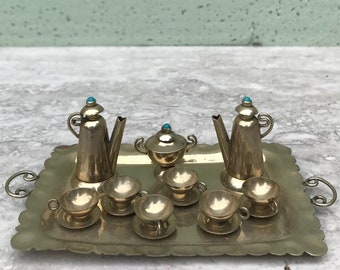 May include: A miniature gold-colored tea set on a decorative tray. The set includes two teapots, a sugar bowl, six teacups with saucers, and a tray with handles. Each piece is detailed with a turquoise stone on top.
