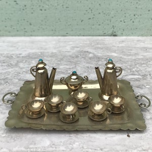 May include: A miniature gold-colored tea set on a decorative tray. The set includes two teapots, a sugar bowl, six teacups with saucers, and a tray with handles. Each piece is detailed with a turquoise stone on top.