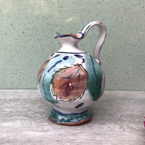 May include: A small, white ceramic pitcher with a brown, green, and blue leaf design. The pitcher has a rounded body and a short, wide spout.