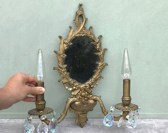 May include: A gold-toned wall sconce with a mirror and two candle-style light fixtures. Each light fixture has a crystal teardrop pendant.