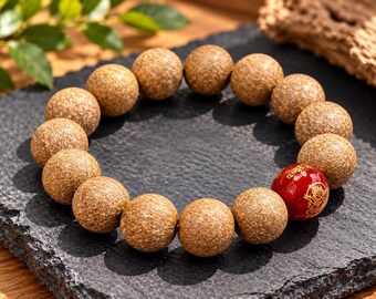 Puede incluir: Una pulsera de cuentas con numerosas cuentas de madera redondas, texturizadas y de color marrón claro dispuestas en círculo. Una cuenta es una esfera roja lisa y brillante decorada con intrincados dibujos y letras doradas.