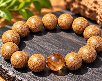 Puede incluir: Una pulsera de cuentas con numerosas cuentas de madera redondas, texturizadas y de color marrón claro, y una cuenta central lisa, translúcida y de color ámbar. Las cuentas están dispuestas en círculo, mostrando un contraste entre la madera mate moteada y la piedra brillante.