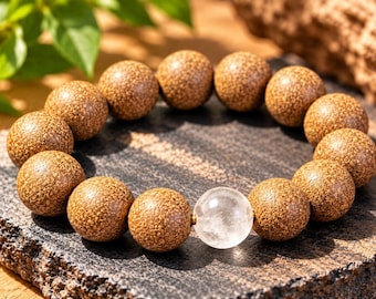 Puede incluir: Una pulsera de cuentas que presenta trece cuentas esféricas de madera marrón con una superficie texturizada y moteada, y una cuenta de cristal esférica, transparente y lisa, colocada como punto focal en la disposición circular.