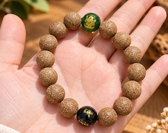 Puede incluir: Una pulsera de cuentas con numerosas esferas marrones texturizadas, una cuenta verde lisa con una ilustración dorada de una deidad y una cuenta negra lisa con letras doradas en caligrafía.