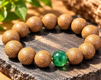 Puede incluir: Una pulsera de cuentas que presenta numerosas cuentas de madera marrón redondas y texturizadas, junto con una cuenta de piedra preciosa verde, lisa y translúcida. Las cuentas están dispuestas en un patrón circular, destacando el contraste entre la madera mate y la piedra brillante.