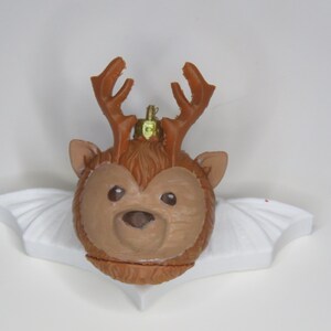 May include: A brown reindeer ornament with a light brown face and antlers. The ornament has a gold-colored loop for hanging and is resting on a white, wing-shaped base. The reindeer has large, dark eyes.