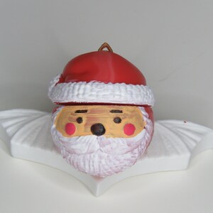 May include: A Santa Claus ornament with a red hat and white trim, a painted face, and a white beard. The ornament is on a white, fan-shaped base, perfect for holiday decor.