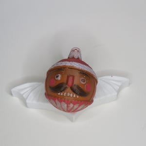 May include: A vintage Christmas ornament shaped like a nutcracker's head, featuring a brown face, black mustache, and red cheeks. The ornament has a red and white hat and is resting on a white, wing-shaped base.