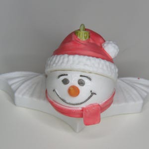 May include: A ceramic snowman Christmas ornament. The snowman has a red hat, scarf, and a carrot nose. The ornament is white and sits on a white, wing-shaped base. A festive holiday decoration.