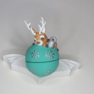 May include: A teal Christmas ornament with silver snowflake designs and a small brown and white reindeer figurine on top. The ornament sits on a white decorative base, creating a festive holiday decoration.