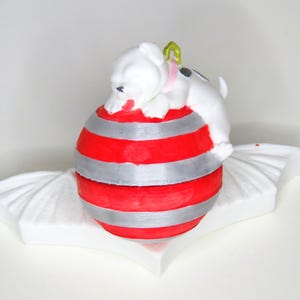 May include: A white ceramic puppy figurine with black spots and a green collar rests on a red and silver striped ball. The ball sits on a white, wing-shaped base. The figurine has a pink tongue.
