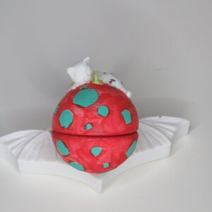 May include: A whimsical, hand-painted ceramic trinket box. The box is a vibrant red with teal polka dots, topped with a white cat figurine. It rests on a white, wing-shaped base, creating a playful and decorative piece.