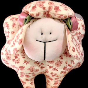 Puede incluir: Un peluche de oveja con cuerpo color crema y estampado floral en tonos rosas y rojos. La oveja tiene una cara blanca con rasgos negros cosidos y mejillas rosas. Un lazo rosa y una cinta verde adornan su cabeza.