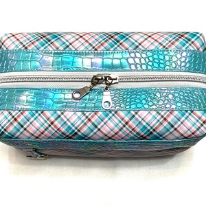 May include: A rectangular cosmetic bag with a plaid pattern in pink, white, and teal. The bag features a teal, faux-crocodile skin band around the middle and a silver zipper. The bag is designed for storing makeup and other beauty products.