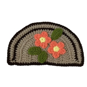 Puede incluir: Una alfombra de crochet en forma de semicírculo con un borde marrón y un centro gris. La alfombra tiene dos flores rosas y amarillas con hojas verdes.