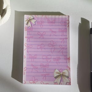Könnte beinhalten: Ein rosa Notizblock mit Schleifenmuster und linierten Seiten. Ein silberner Stift und ein weißer Kerzenhalter mit einer Kerze befinden sich neben dem Notizblock. Ein Buch mit dem Titel "Mona Lisa" ist im Hintergrund.