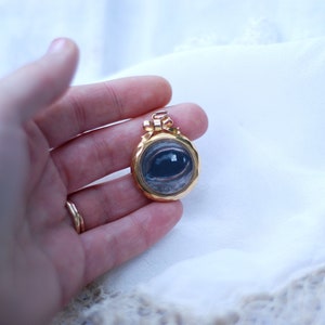 May include: A gold locket pendant with a painted eye design. The eye is blue with a brown iris and a white sclera.