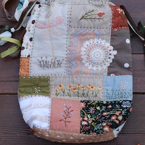 Peut inclure: Un sac à cordon patchwork avec des broderies variées. Le sac présente des carrés de tissu de différentes couleurs, dont le beige, le vert et le marron. La broderie comprend des fleurs, une libellule et un napperon. Le sac est noué avec un ruban.